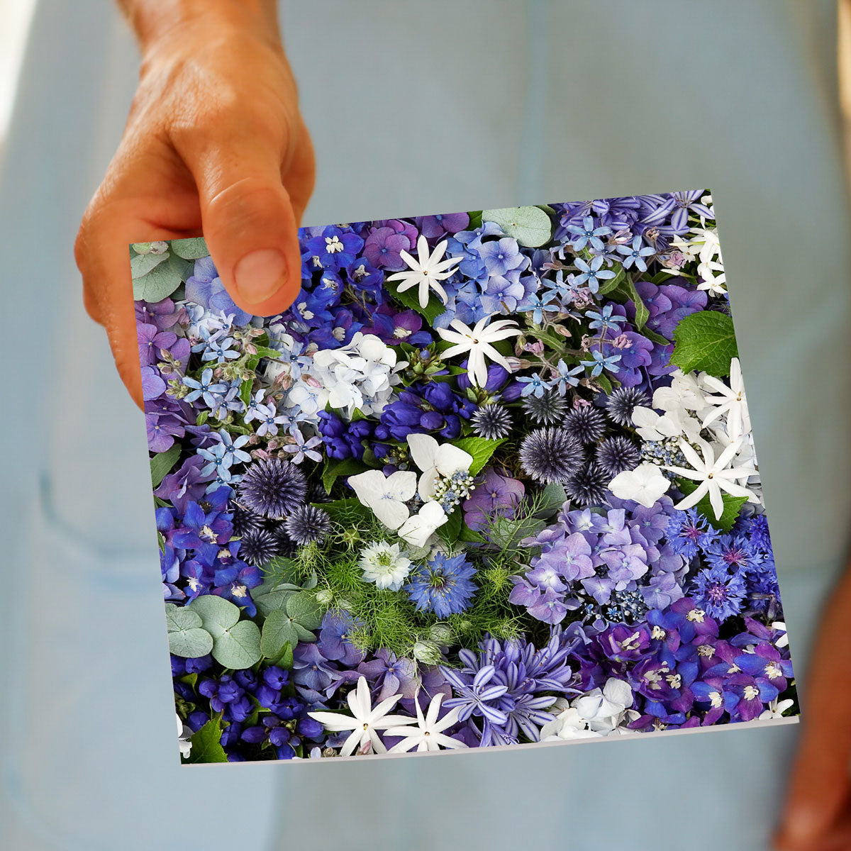 A beautiful greeting card with a blue and white floral arrangement & green leaves. This card is being held by hand.Size 142 x 142mm. Found at Golly Gosh Creations.