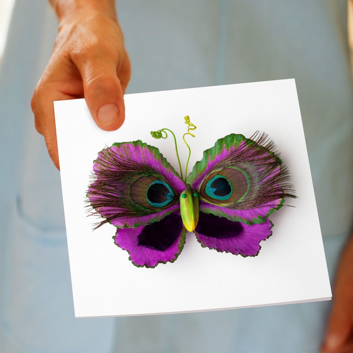 A person holding a vibrant butterfly greeting card. Created from cabbage leaves and peacock feathers This quality square greeting card is found at Golly Gosh Creations.