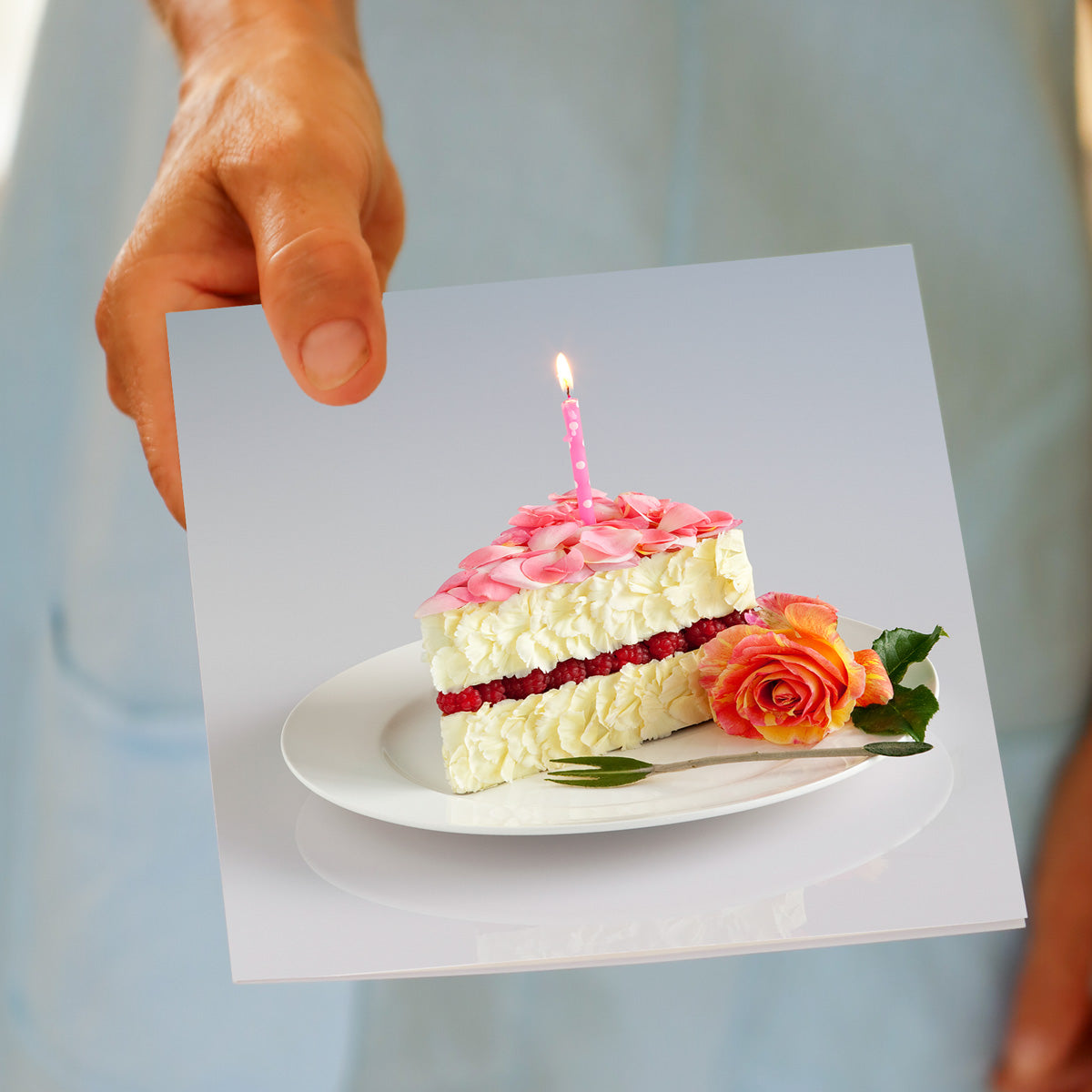 Hand displaying Cake Slice birthday card with floral cake featuring pink rose petal topping, white flower layers, red bloom filling, and glowing birthday candle.