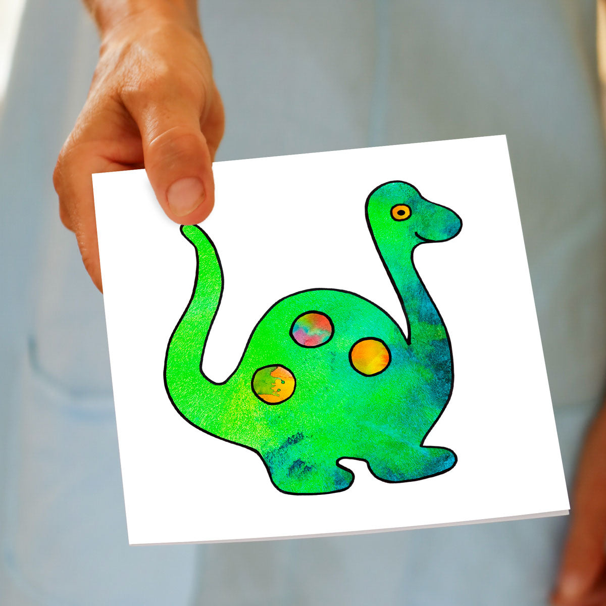 A greeting card, being held by hand.
With a colourful kids drawing of a dinosaur on a white background. A quality square greeting card found at Golly Gosh Creations.