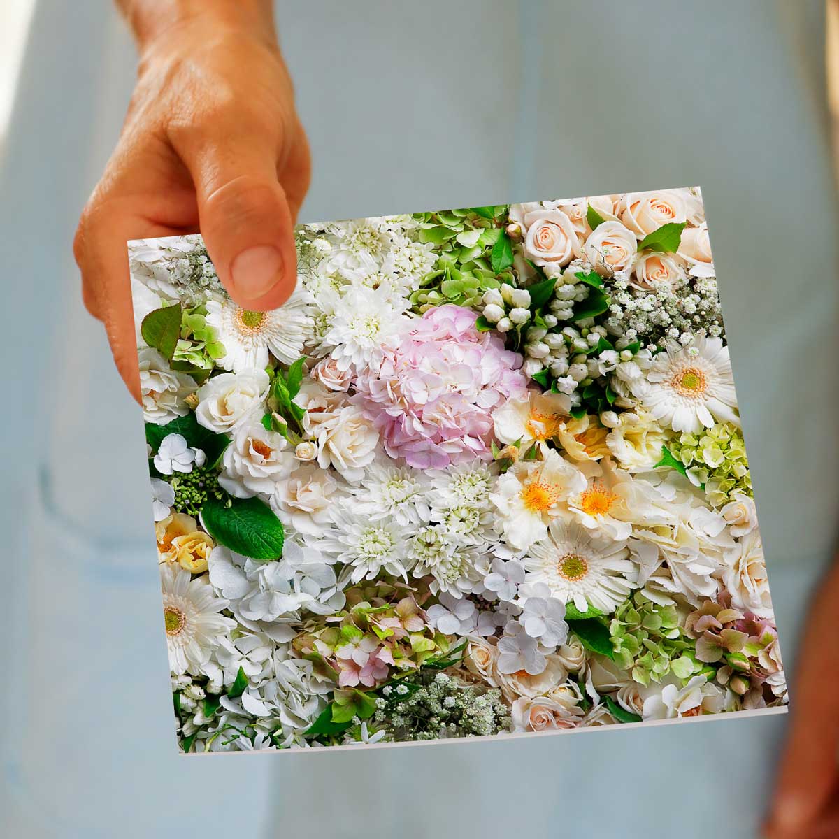 A hand holding our greeting card featuring a floral arrangement of mostly white flowers, accompanied by soft pink and green hydrangeas and apricot coloured roses. Size 142 x 142mm. A quality greeting card found at Golly Gosh Creations.