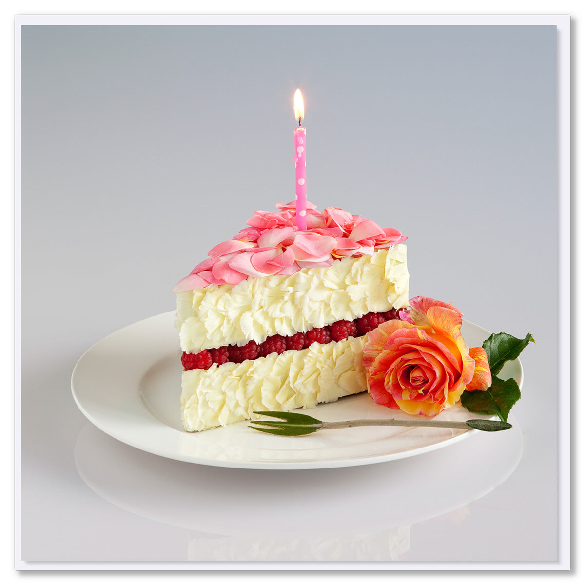 Floral cake slice greeting card made of pink rose petals, white blooms, and red raspberries with lit birthday candle on white plate, coral rose garnish